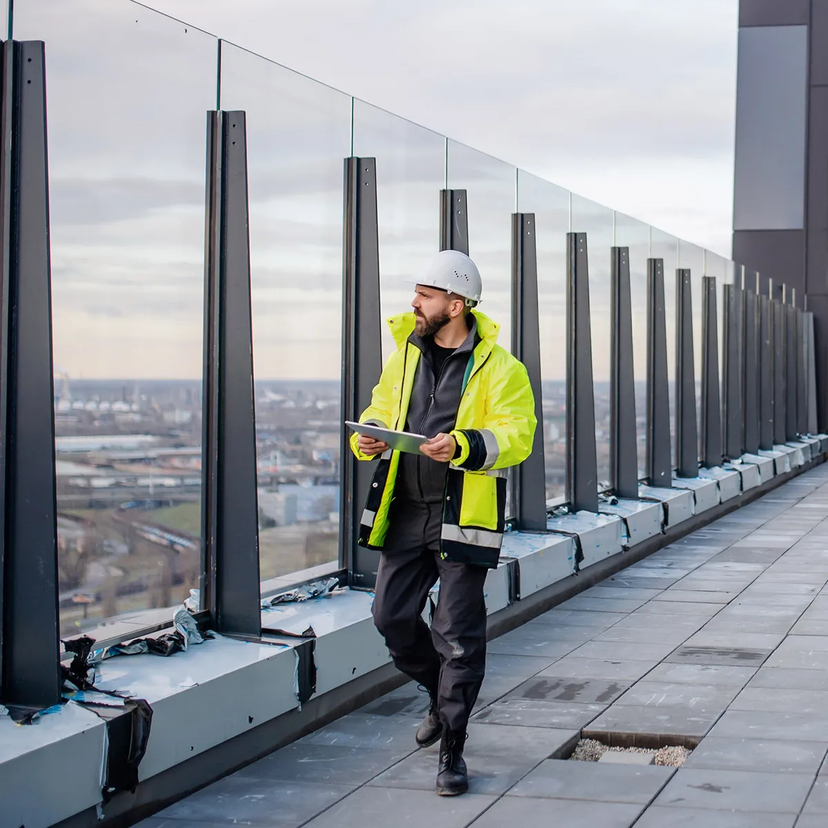 Bouwinspecteur met veiligheidshelm en gele jas die een gebouw buiten inspecteert met een tablet in de hand.